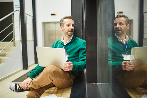 Positive mature conference participant sitting on bench with laptop on his laps and looking through big office window