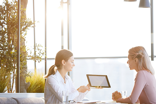 Content young attractive businesswoman sitting at table in restaurant and using tablet while sharing marketing strategy with colleague