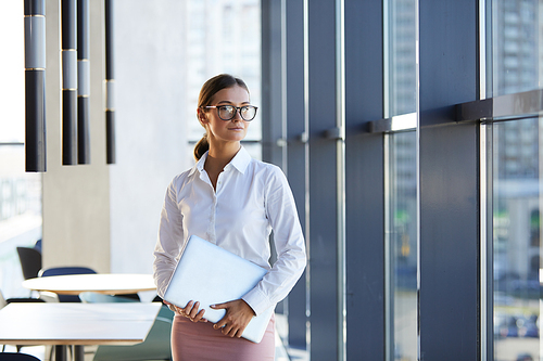 Portrait of content attractive businesswoman in white blouse standing in contemporary office lobby and holding laptop