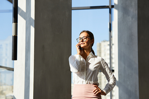 Content purposeful young business lady in glasses standing in modern corridor and talking by phone