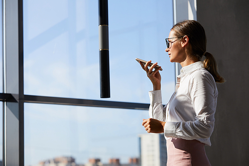 Confident young businesswoman with ponytail standing at window and sending voice message to colleague