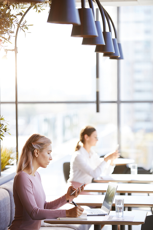 Busy young female freelance administrator in sweater sitting at table and working with digital devices