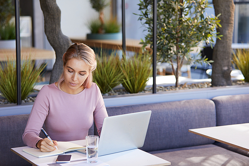 Concentrated young business lady sitting at table in modern cafe and making notes in organizer while planning meetings for day