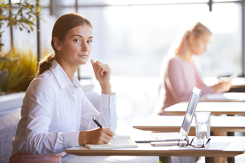 Portrait of serious attractive young businesswoman in formal blouse sitting at table in cafe and preparing for annual meeting