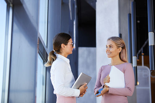 Positive attractive female colleagues in stylish outfits standing at window in corridor and talking about work