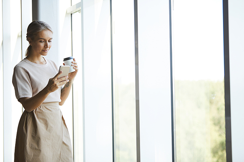 Content attractive blond woman in skirt standing by window and reading message on phone while drinking coffee