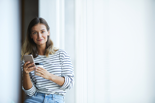 Portrait of smiling beautiful hipster designer in stripped sweater drinking takeout coffee and using smartphone