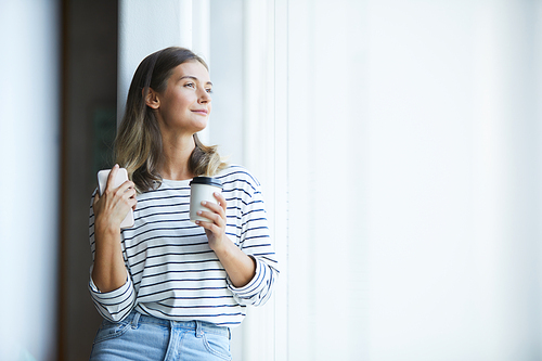 Content dreamy young lady in casual outfit holding smartphone and coffee cup while looking out window