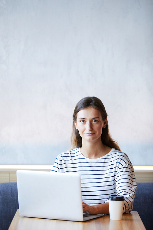 Portrait of content confident young freelancer in black and white stripped sweater sitting at table and working with laptop in cafe with wifi