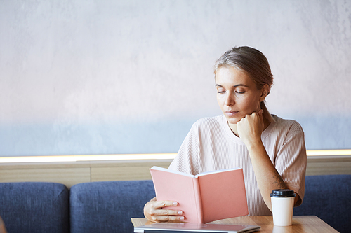 Serious pensive young woman with beautiful color of hair sitting at table and enjoying reading in cafe