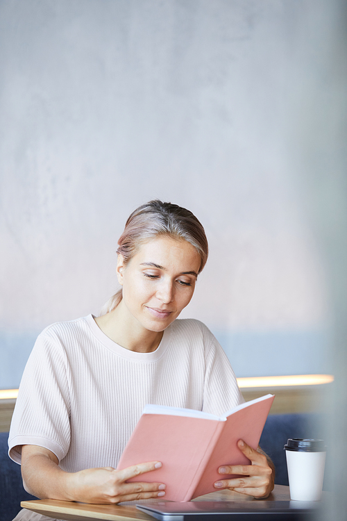 Content curious young lady sitting at table with coffee cup and spending time with interesting book