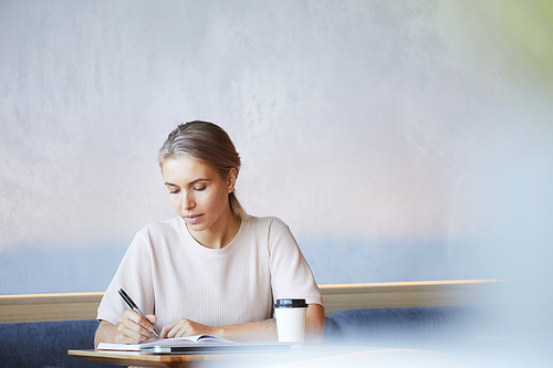 Serious concentrated blond lady in casual sweater sitting at table and making notes in diary while working in cafe