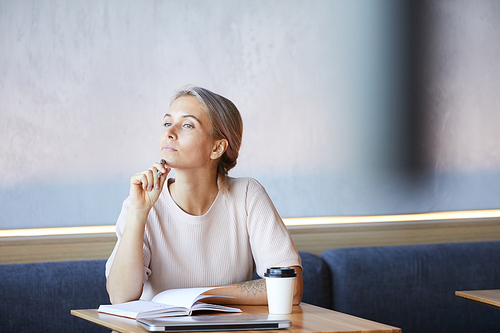 Serious young businesswoman with tattoo on arm sitting at table in coffee shop and thinking of new creative ideas