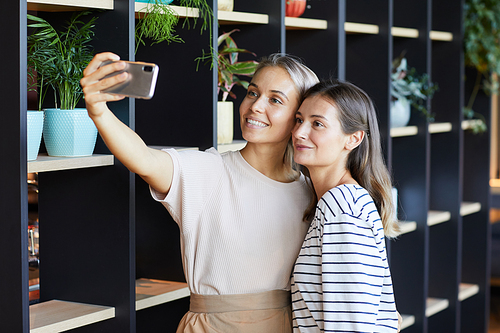 Smiling attractive young woman with pretty toothy smile taking selfie with friend in modern cafe