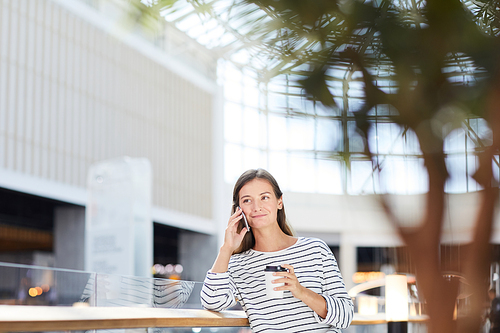 Content confident attractive young woman in stripped sweater leaning on railing while talking by phone in shopping mall