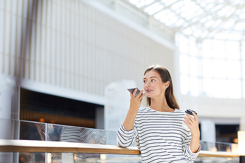 Content confident young woman in casual outfit leaning on railing and sending voice message to colleague while having coffee break