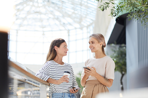 Cheerful attractive young women in casual outfits standing in shopping mall and using smartphones while chatting with each other