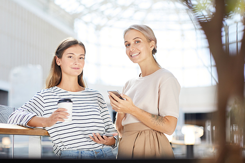 Portrait of happy beautiful young female friends in casual outfits standing in modern lobby and using gadgets