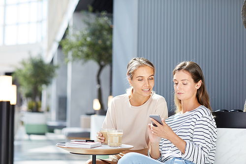 Positive modern girls in casual clothing sitting on sofa and drinking coffee while surfing social medias together