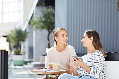 Smiling pretty women in casual outfits sitting at food court of shopping mall and using smartphone while chatting with each other