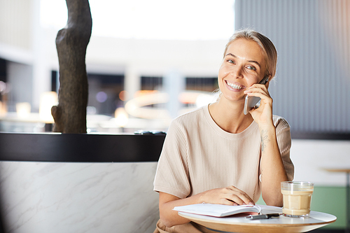 Portrait of happy attractive young woman working as freelance telemarketer remotely talking on phone in cafe