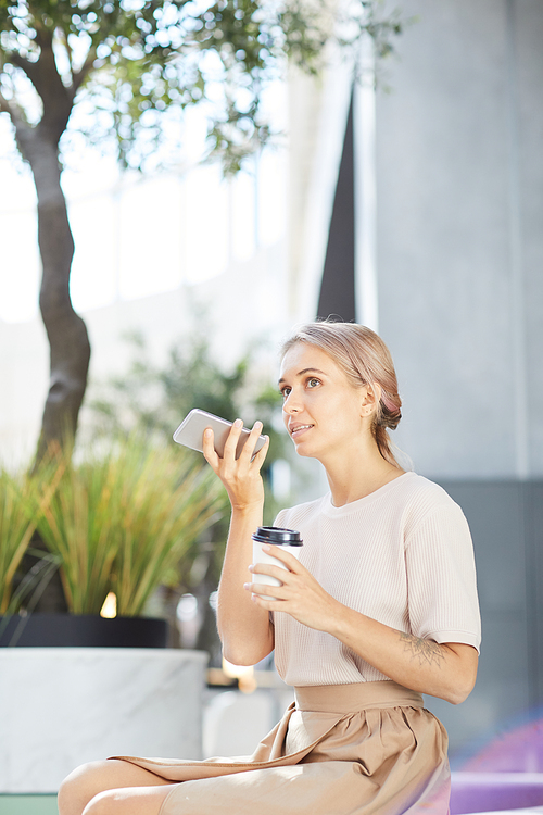 Pensive young lady in stylish clothes sitting on bench in shopping mall and answering friend using instant audio message