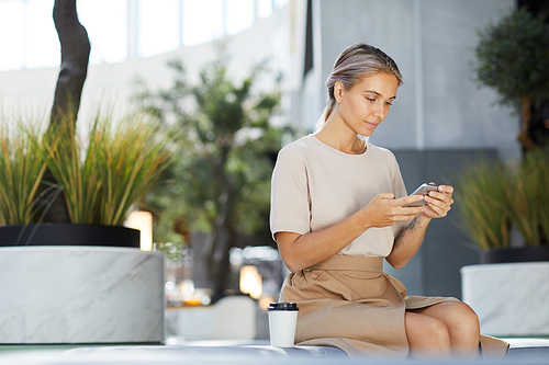 Content pensive young woman with tattoo on arm sitting on bench in shopping center and checking messenger on phone