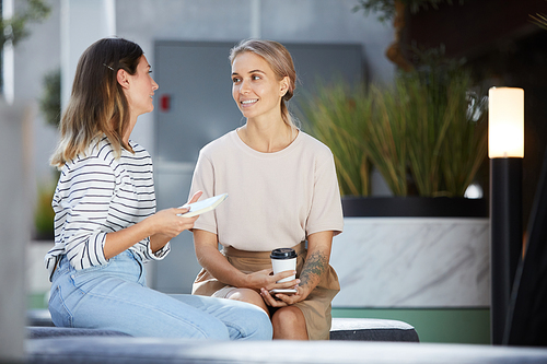 Positive pretty young ladies in casual clothing sitting on bench in contemporary office lobby and talking during coffee break