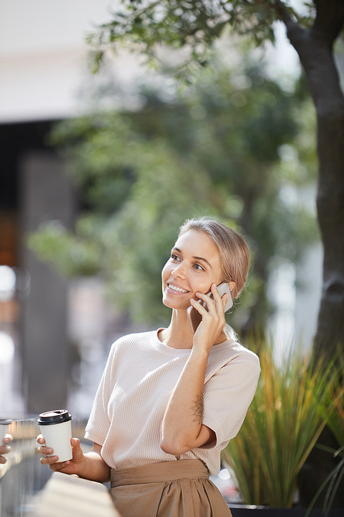 Smiling confident female manager with tattoo standing in city park and drinking coffee while having phone conversation