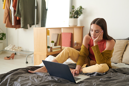 Tired young brunette woman in cardigan sitting on bed and leaning head on hand while using laptop and doing boring job at home