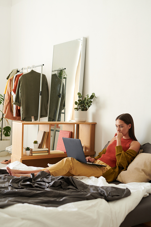 Thoughtful young woman in cardigan lying on comfortable bed and using laptop while working from home