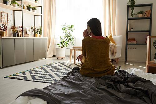 Rear view of brunette woman in cardigan sitting on bed and calling close friend by cellphone in studio apartment