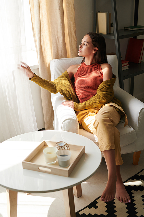 Serious beautiful young woman in mustard-colored cardigan sitting in armchair and adjusting curtains while waiting for husband at home