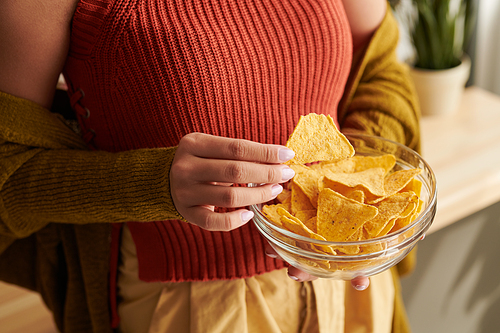 Close-up of unrecognizable woman in bright top and cardigan eating nachos from bowl at home, unhealthy food concept
