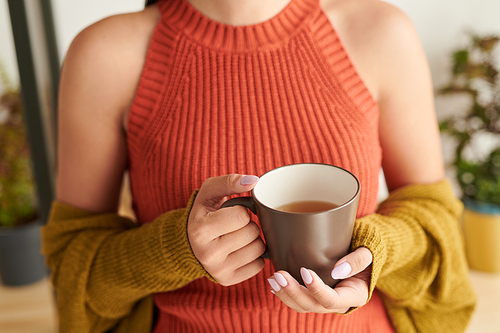 Close-up of unrecognizable girl in orange top and cardigan holding mug of tea while enjoying morning at home