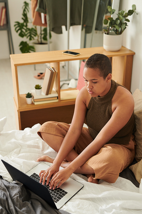 Concentrated young African-American woman sitting with crossed legs on bed and typing on laptop while filling report at home