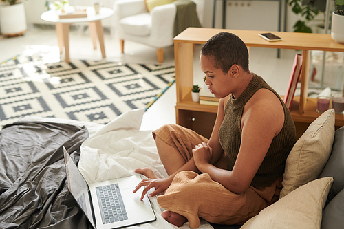 Focused young black woman with buzz cut sitting on bed and using laptop touchpad in studio flat