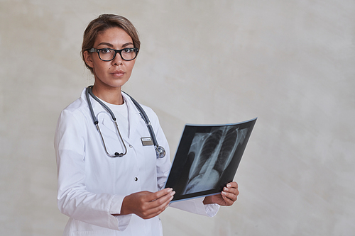 Studio portrait of attractive female doctor wearing eyeglasses and white coat holding chest X-ray shot looking at camera