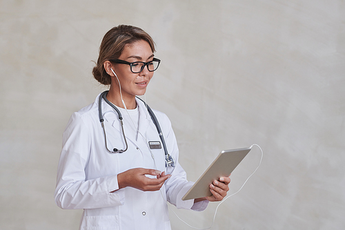 Horizontal medium studio portrait of modern female doctor wearing eyeglasses and earphones using digital tablet for online appointment