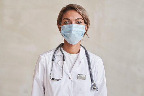 Horizontal medium close-up portrait of modern young adult doctor wearing protective mask looking at camera