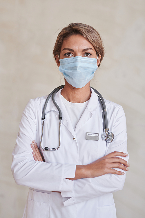 Vertical medium portrait of confident young adult therapist wearing protective mask standing with arms crossed looking at camera