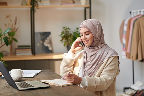 Beautiful Muslim businesswoman wearing hijab sitting at desk discussing work issues on phone and making notes