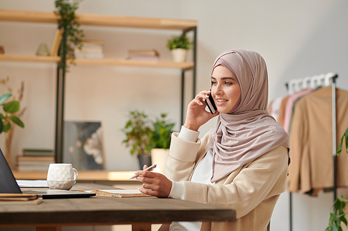 Modern Muslim woman working as fashion designer sitting at desk in cozy office room talking on phone