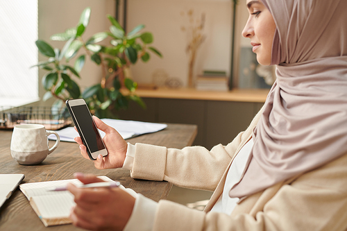 Modern Muslim businesswoman wearing headscarf sitting at office desk checking messages on her smartphone