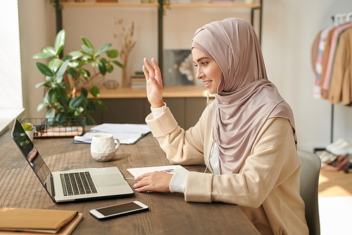 Cheerful young Muslim woman wearing pale pink headscarf sitting in front of laptop starting online meeting with greeting her colleagues