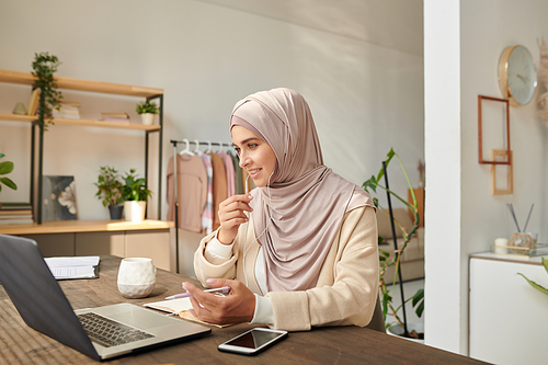 Modern Muslim woman wearing headscarf working at home taking part in online meeting discussing something with her coworkers