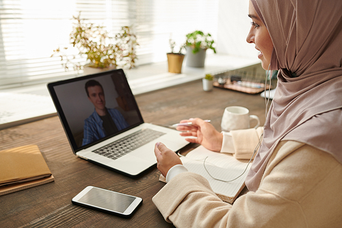 Stylish Muslim woman wearing hijab discussing new business project issues with her male colleague using online meeting application