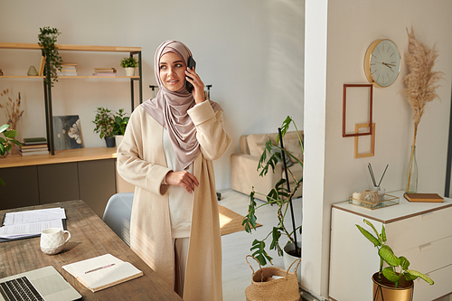 Horizontal shot of young Muslim woman wearing headscarf standing at desk talking on phone with her friend looking away, copy space