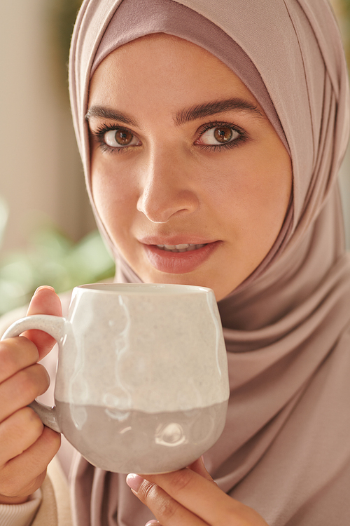 Vertical close-up portrait of young adult attractive Middle Eastern woman wearing stylish pastel pink hijab drinking tea looking at camera