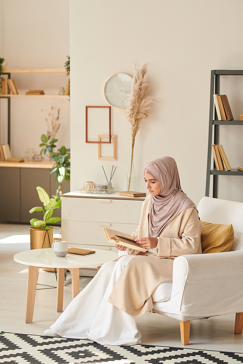 Vertical shot of modern young Muslim woman wearing stylish outfit with hijab sitting in armchair reading book in living room